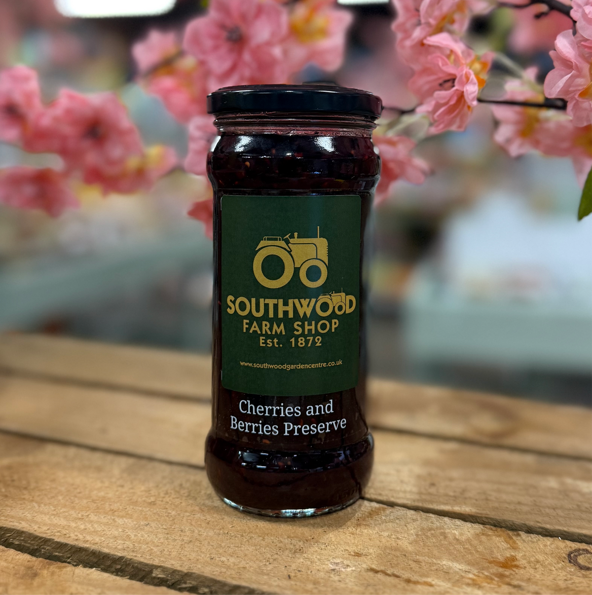 A jar of Southwood Farmshop Cherries & Berries Preserve on a wooden surface with pink flowers in the background.