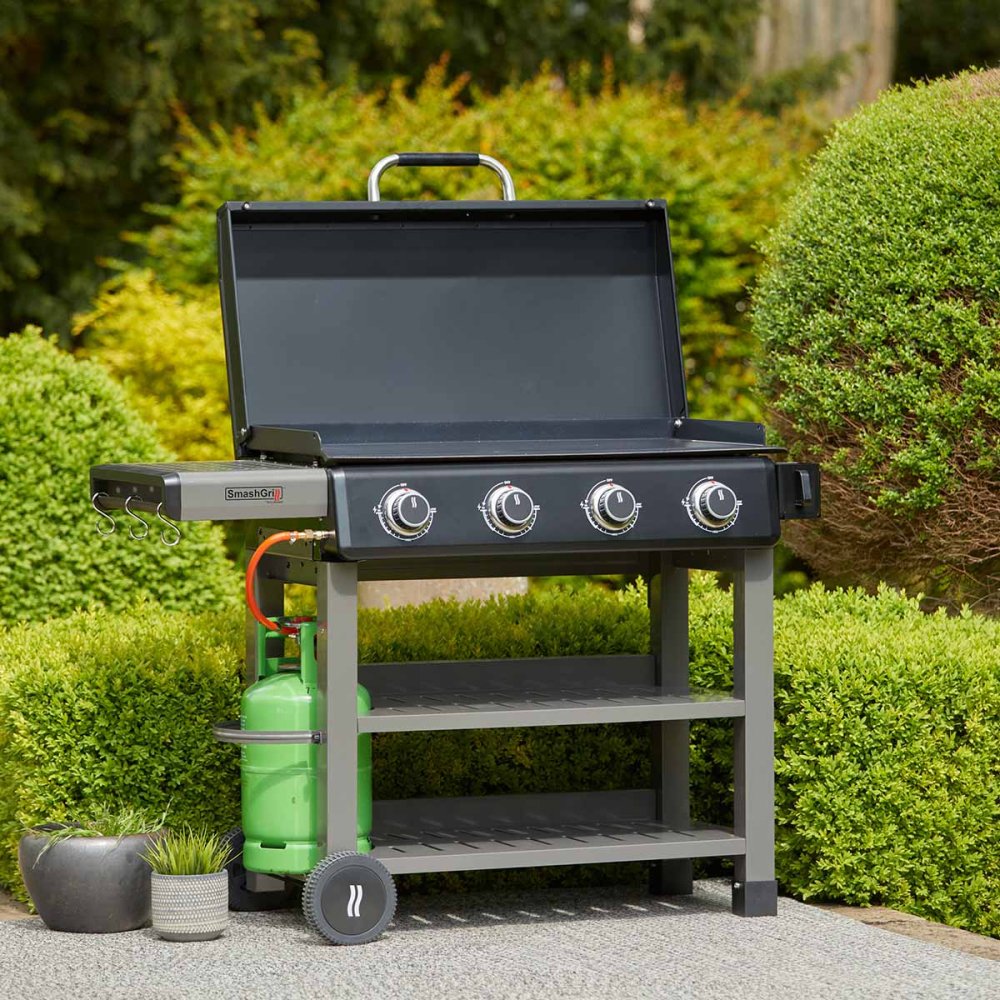 A black Grillstream SmashGrill 4 Burner Flat Plate Gas BBQ / Barbecue with U-shaped burners, integrated Piezo ignition, and a removable fat cup, set in an outdoor garden setting.